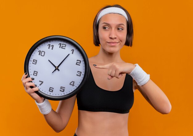 Giovane donna fitness in abiti sportivi con fascia tenendo l'orologio da parete puntato con il dito con un sorriso fiducioso sul viso in piedi sopra la parete arancione