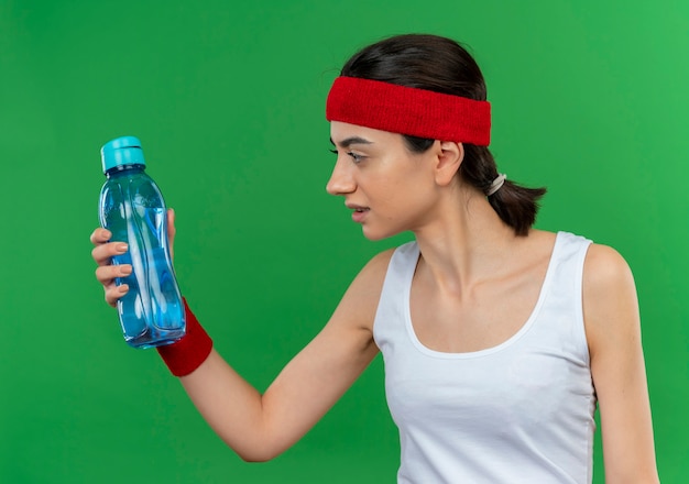 Giovane donna fitness in abiti sportivi con fascia tenendo la bottiglia di acqua guardandolo confuso in piedi sopra la parete verde