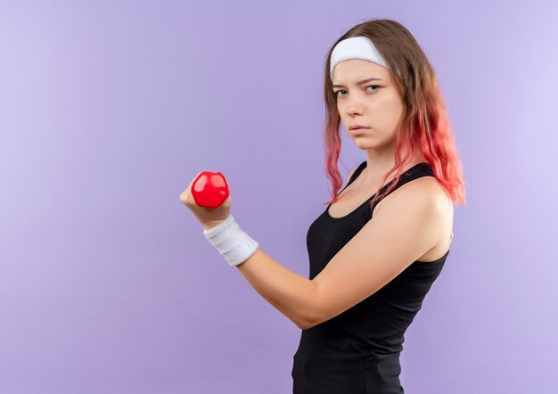 Giovane donna fitness in abiti sportivi facendo esercizi con manubri con faccia seria in piedi sopra la parete viola