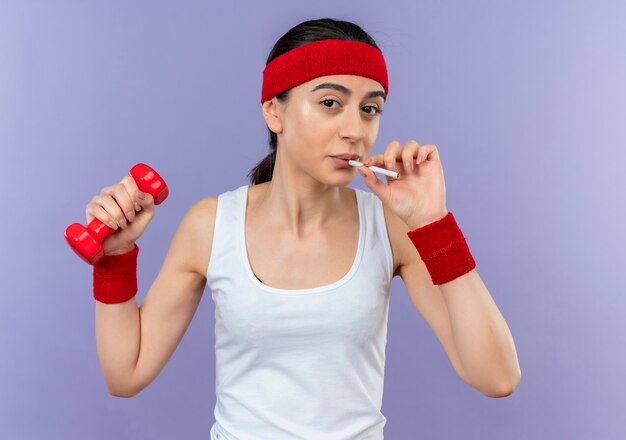 Giovane donna fitness in abiti sportivi tenendo il manubrio con la sigaretta andando a fumare in piedi sopra la parete viola