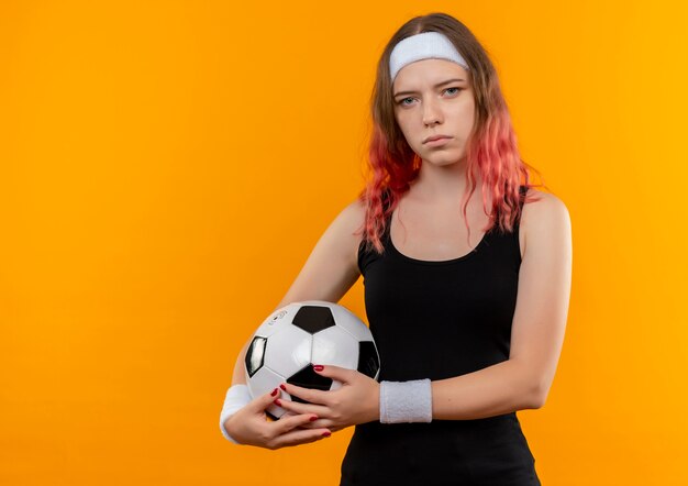 Giovane donna fitness in abiti sportivi tenendo il pallone da calcio nelle mani con la faccia seria in piedi sopra la parete arancione
