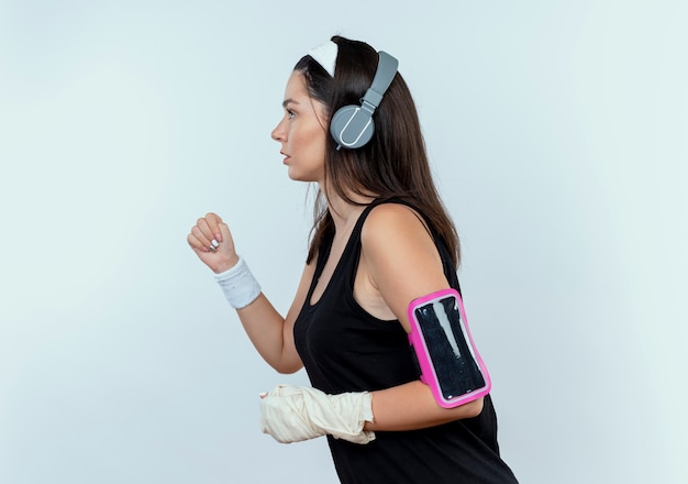 Giovane donna fitness in archetto con cuffie e fascia da braccio dello smartphone che lavora fuori in piedi su sfondo bianco