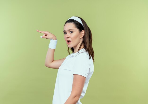 giovane donna fitness in fascia cercando confuso puntando con il dito sul lato in piedi sopra la parete chiara