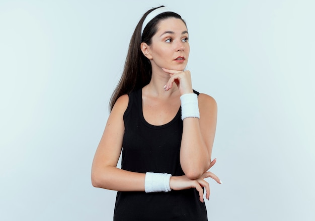Giovane donna fitness in fascia cercando da parte con la mano sul mento pensando in piedi su sfondo bianco
