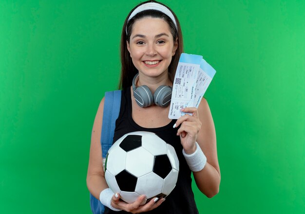 Giovane donna fitness in fascia con le cuffie e lo zaino in possesso di pallone da calcio e biglietti aerei guardando la fotocamera sorridente con la faccia felice in piedi su sfondo verde