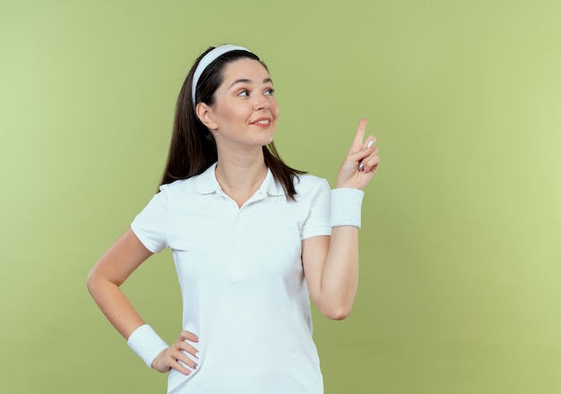 giovane donna fitness in fascia guardando da parte che punta con il dito a qualcosa di sorridente fiducioso in piedi sopra il muro di luce