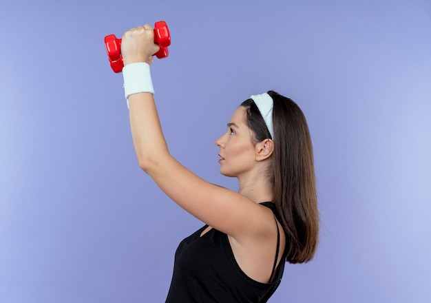 Giovane donna fitness in fascia lavorando con manubri cercando fiducioso in piedi oltre il muro blu