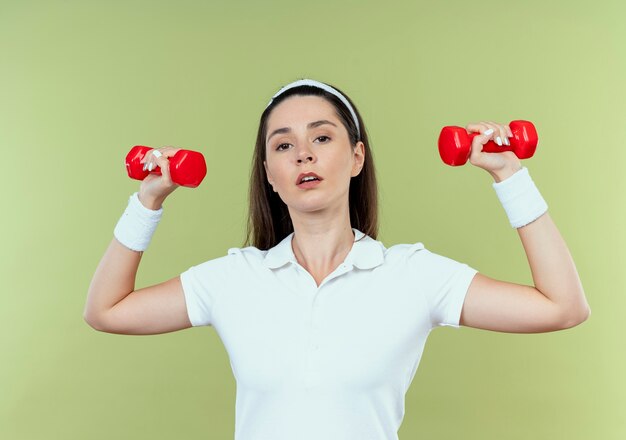 Giovane donna fitness in fascia lavorando con manubri cercando fiducioso in piedi sopra la parete leggera