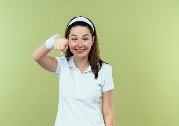 Giovane donna fitness in fascia sorridendo allegramente puntando il dito alla fotocamera in piedi su sfondo chiaro