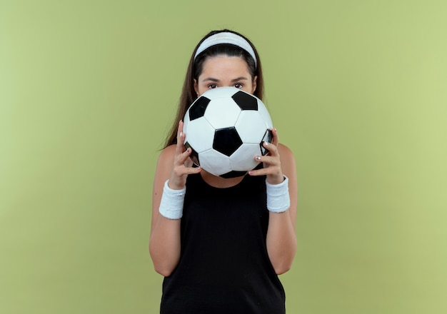 Giovane donna fitness in fascia tenendo il pallone da calcio che nasconde il viso dietro la palla che dà una occhiata in piedi su sfondo chiaro