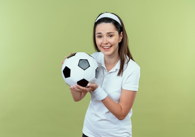 giovane donna fitness in fascia tenendo il pallone da calcio sorridendo allegramente in piedi sopra il muro di luce