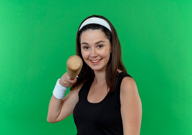 giovane donna fitness in fascia tenendo la mazza da baseball che punta sorridente in piedi sopra il muro verde