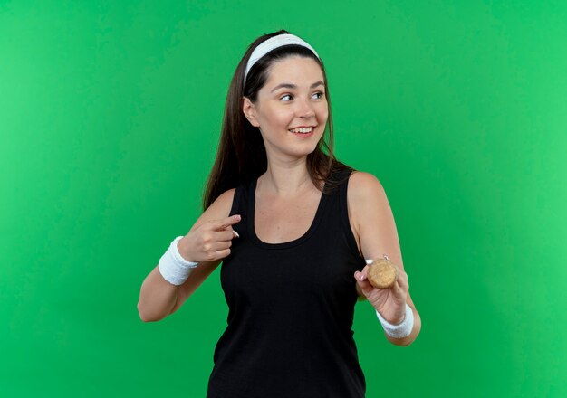 Giovane donna fitness in fascia tenendo la mazza da baseball guardando da parte sorridente con la faccia felice che punta con il dito a lato in piedi su sfondo verde