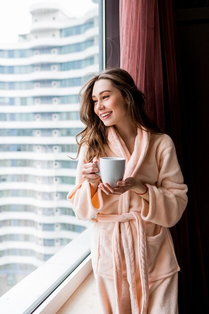 Giovane donna fresca in accappatoio rosa tenero beve il tè, guardando fuori dalla finestra.