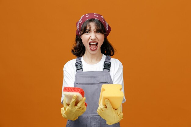 Giovane donna furiosa addetta alle pulizie che indossa guanti di gomma uniformi e bandana che tiene spugne guardando la fotocamera che grida ad alta voce isolata su sfondo arancione