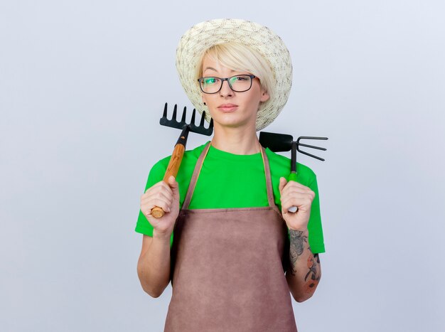 Giovane donna giardiniere con i capelli corti in grembiule e cappello con zappa e mini rastrello con faccia seria