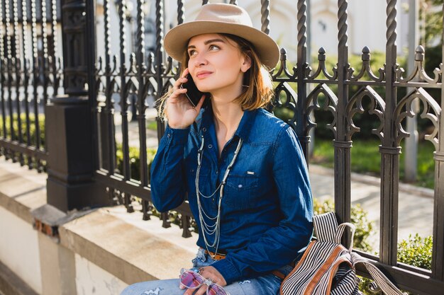 Giovane donna graziosa alla moda che sorride e che parla sul suo telefono, vestito in jeans e camicia di jeans