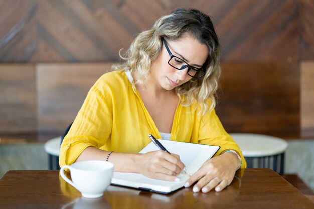 Giovane donna graziosa che beve caffè e piani di scrittura per il giorno