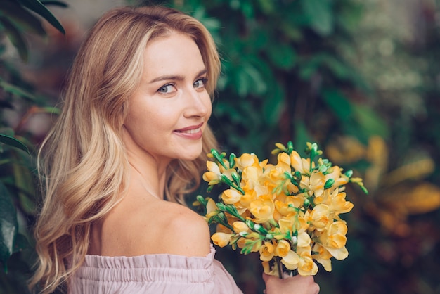 Giovane donna graziosa che esamina macchina fotografica che tiene i bei fiori gialli