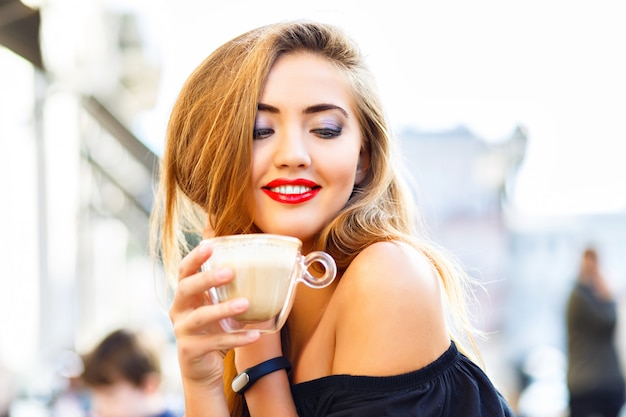 Giovane donna graziosa che gode, che beve tazza di cappuccino, latte, caffè in caffè di strada di mattina.