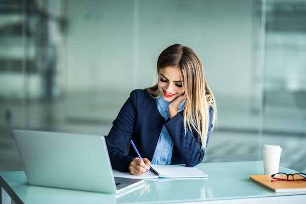 Giovane donna graziosa che lavora con il laptop e che prende appunti su un desktop in ufficio