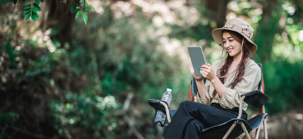 Giovane donna graziosa che si siede sulla sedia e usa la videochiamata del tablet mentre si accampa nello spazio della copia del parco naturale