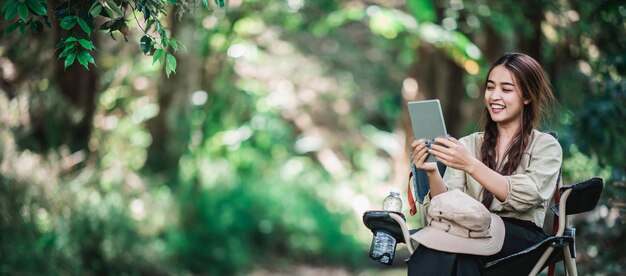 Giovane donna graziosa che si siede sulla sedia e usa la videochiamata del tablet mentre si accampa nello spazio della copia del parco naturale