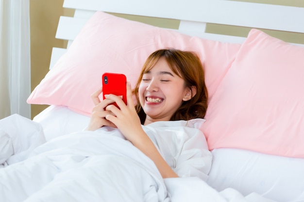 Giovane donna graziosa che si trova sullo smartphone di uso del letto