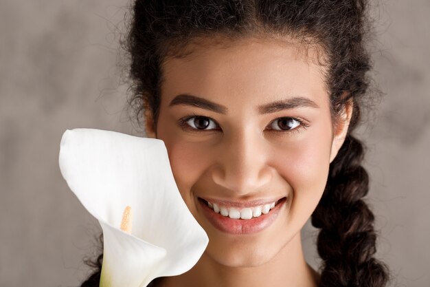 Giovane donna graziosa che sorride, tenendo fiore bianco sopra la parete beige