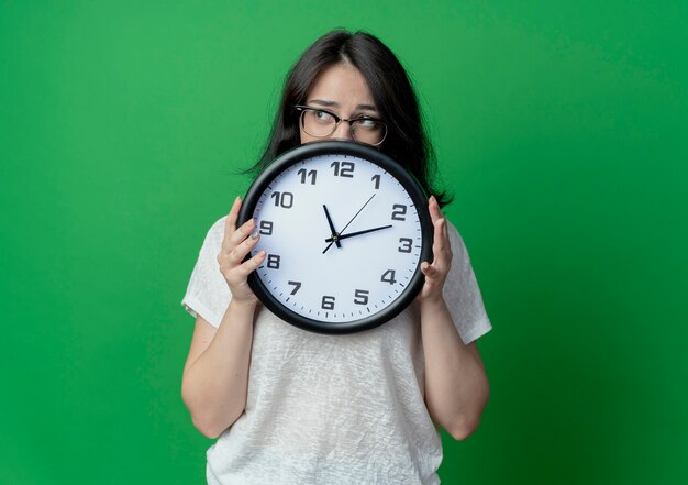 giovane donna graziosa con gli occhiali tenendo l'orologio e guardando il lato da dietro l'orologio