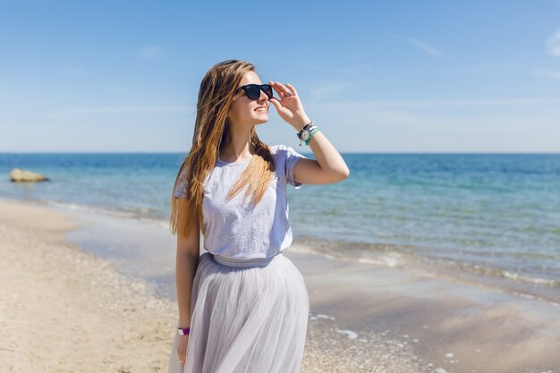 Giovane donna graziosa con i capelli lunghi sta camminando sulla spiaggia vicino al mare