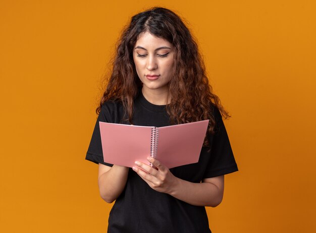 Giovane donna graziosa concentrata che scrive con la matita sul blocco note isolato sulla parete arancione con lo spazio della copia