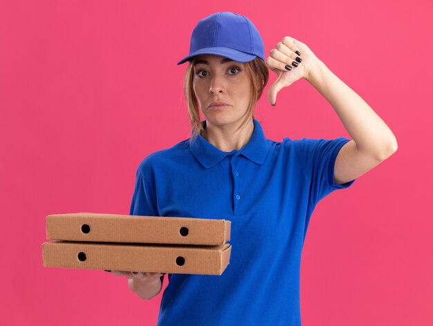 Giovane donna graziosa di consegna delusa in uniforme pollice in giù e tiene scatole per pizza isolate sulla parete rosa