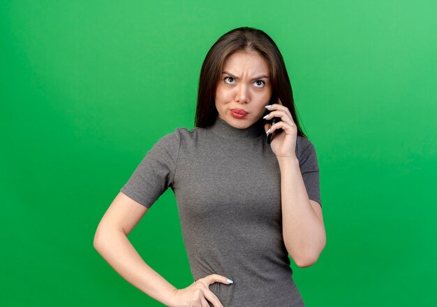 Giovane donna graziosa dispiaciuta che parla sul telefono che mette la mano sulla vita che osserva in su isolato su fondo verde con lo spazio della copia