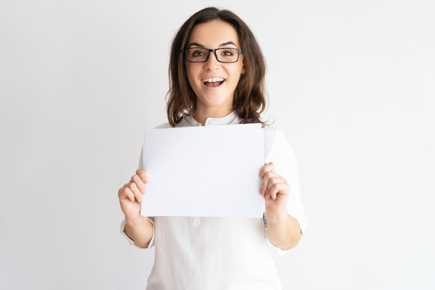 Giovane donna graziosa emozionante che mostra foglio di carta in bianco e che esamina macchina fotografica