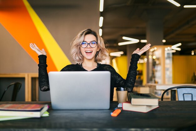 Giovane donna graziosa emozionante felice divertente che si siede al tavolo in camicia nera che lavora al computer portatile in ufficio di co-working, con gli occhiali