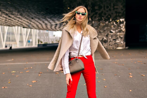 Giovane donna graziosa hipster in posa per strada vicino a moderni centri commerciali, indossando abiti da ufficio alla moda e cappotto di cashmere, inviando un bacio e godendosi una fresca giornata autunnale, colori tonica.