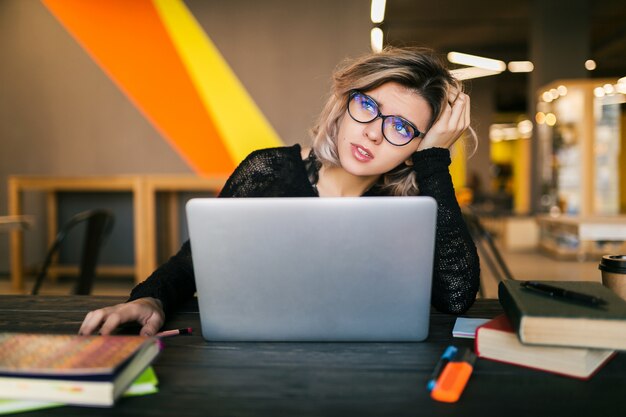 Giovane donna graziosa triste stanca che si siede al tavolo che lavora al computer portatile nell'ufficio di co-working, con gli occhiali, stress sul lavoro, emozione divertente, studente in aula, frustrazione