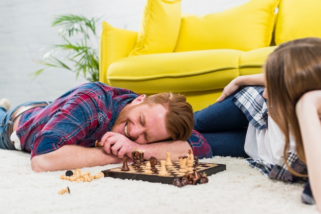 Giovane donna guardando sorridente uomo sdraiato sul tappeto bianco, giocando a scacchi