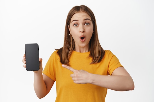 Giovane donna impressionata che punta il dito alla vendita dello schermo dello smartphone, ansimando stupita, mostrando un fantastico affare promozionale sul telefono cellulare, muro bianco.