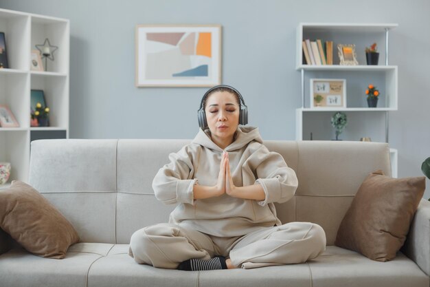 Giovane donna in abiti da casa seduta su un divano a casa interno con le cuffie felice e positivo rilassante meditando facendo gesto di meditazione tenendo i palmi delle mani insieme