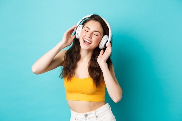 Giovane donna in abiti estivi ascoltando musica, indossando le cuffie e cantando lungo la canzone preferita, ballando con gli auricolari, in piedi su sfondo blu.