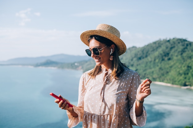 Giovane donna in abito carino estivo, cappello di paglia e occhiali da sole ballando con lo smartphone a portata di mano e ascoltando musica