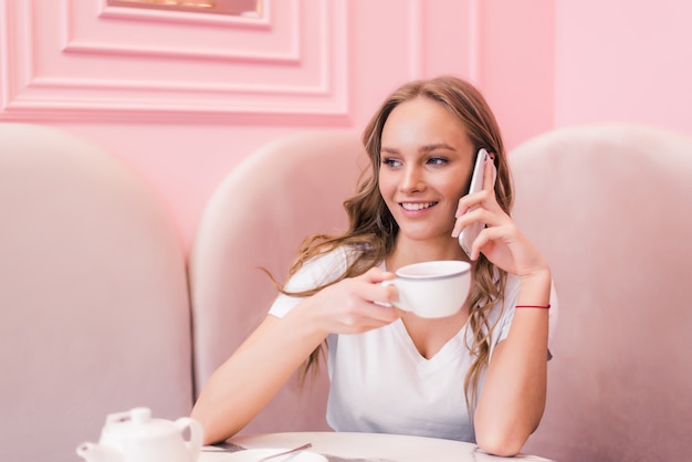 Giovane donna in abito grigio seduto al tavolo in caffè, parlando al cellulare mentre si prendono appunti in taccuino.