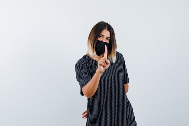 Giovane donna in abito nero, maschera nera che mostra attesa su un gesto minuto, tenendo la mano dietro la vita e guardando felice, vista frontale.