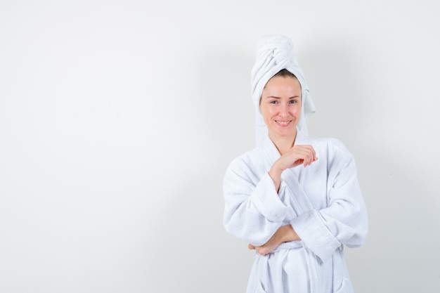 Giovane donna in accappatoio bianco, asciugamano in posa con la mano alzata e guardando allegro, vista frontale.