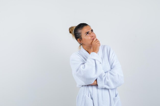 Giovane donna in accappatoio bianco in piedi in posa di pensiero e guardando pensierosa