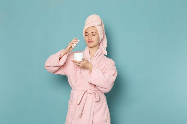 giovane donna in accappatoio rosa dopo la doccia tenendo e usando la crema sul blu