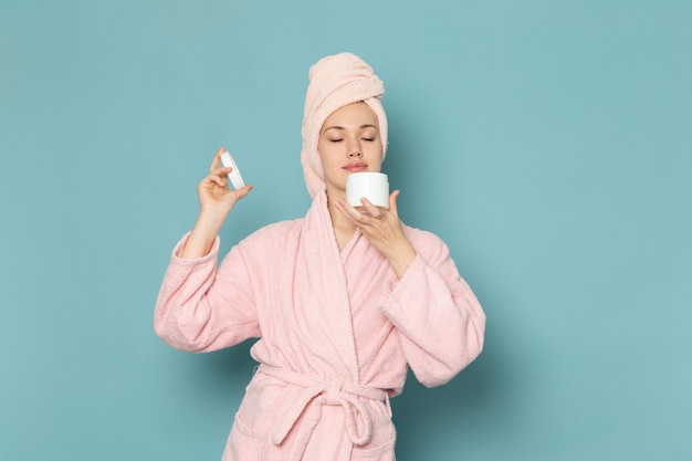 giovane donna in accappatoio rosa dopo la doccia tenendo e usando la crema sul blu