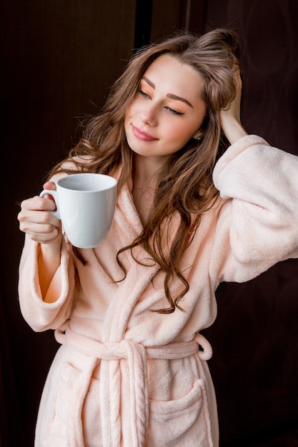 Giovane donna in accappatoio rosa tenero bere il tè e sorridente.